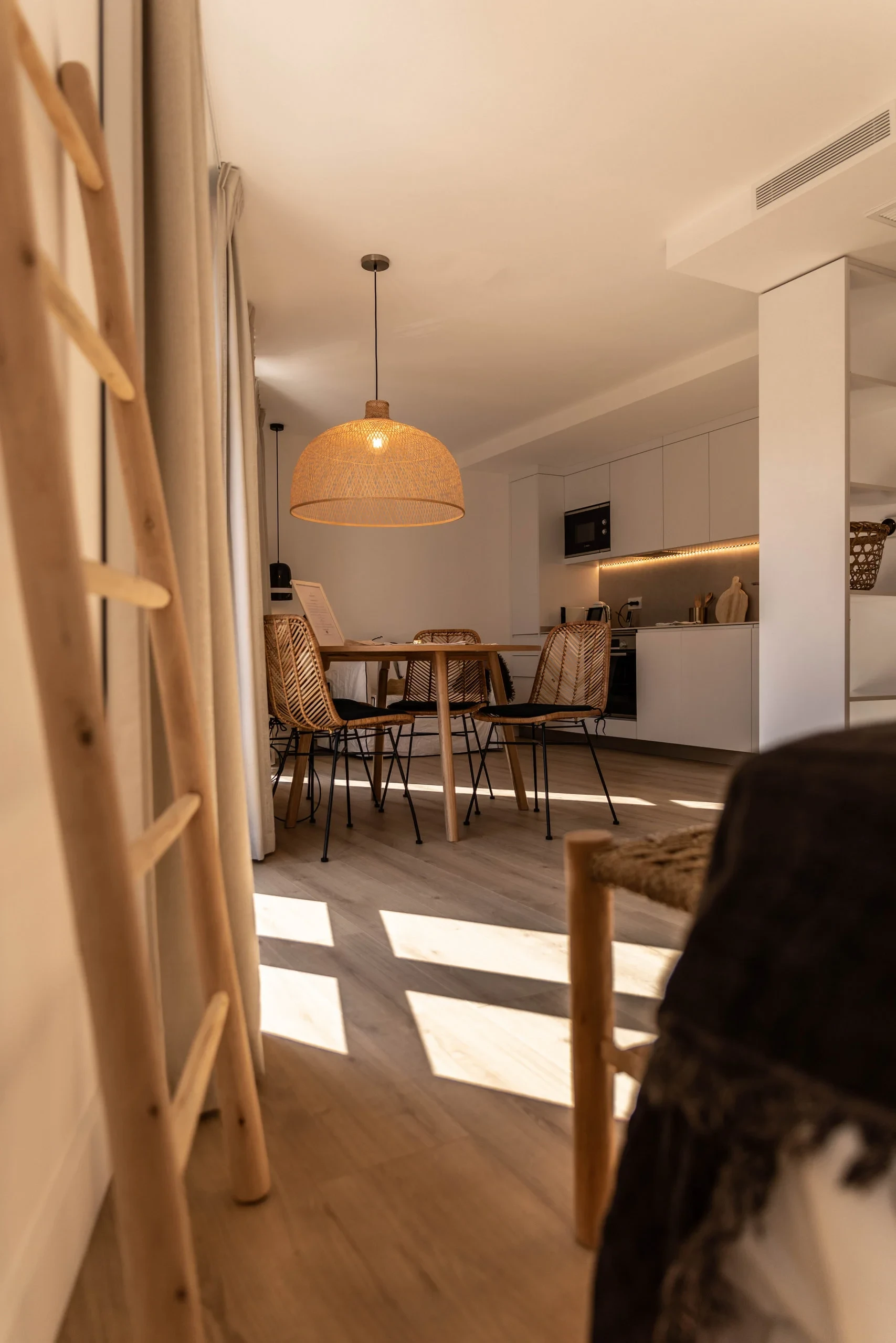Femme avec chapeau de paille assise à une table en bois dans une salle à manger lumineuse - Appartement Vacances Perpignan - Les Suites Collection