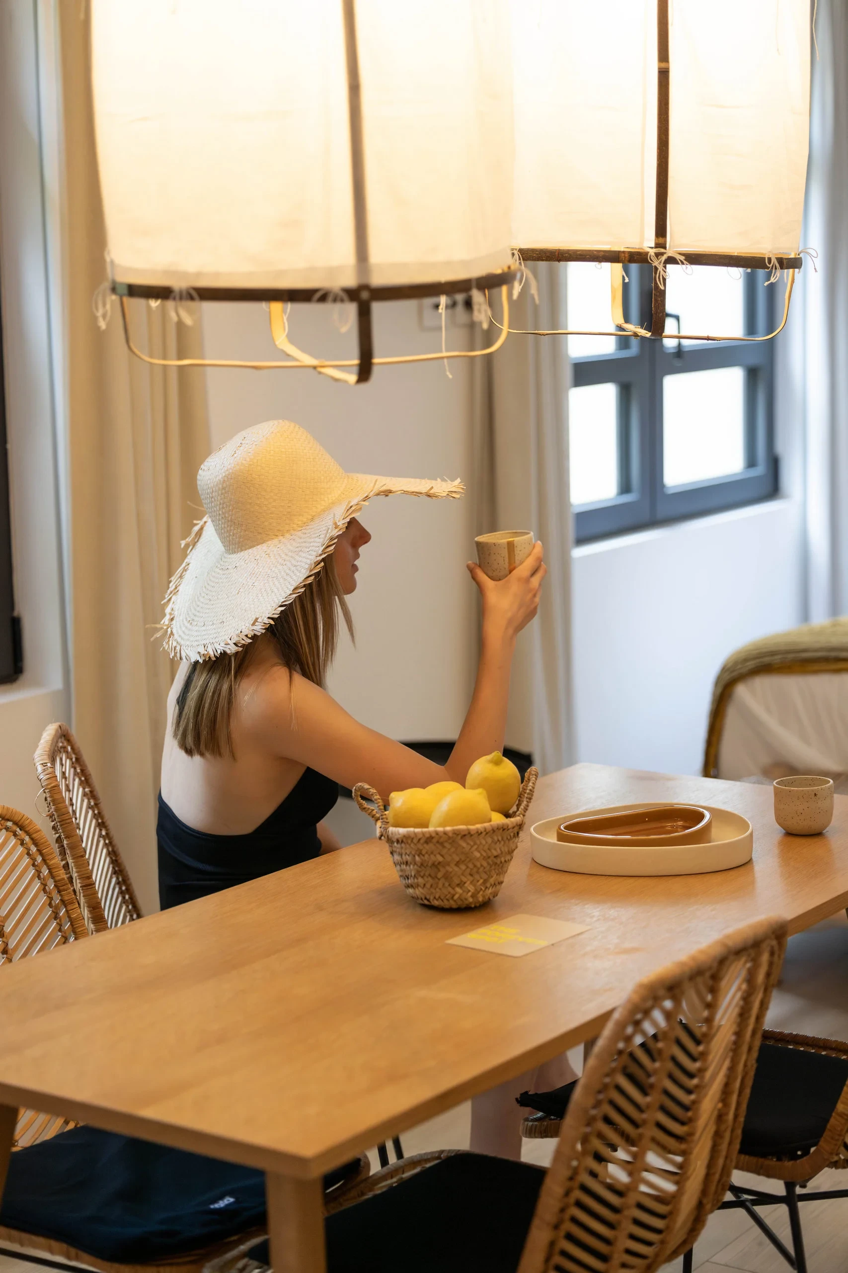 Femme avec chapeau de paille assise à une table en bois dans une salle à manger lumineuse - Appartement Vacances Perpignan - Les Suites Collection