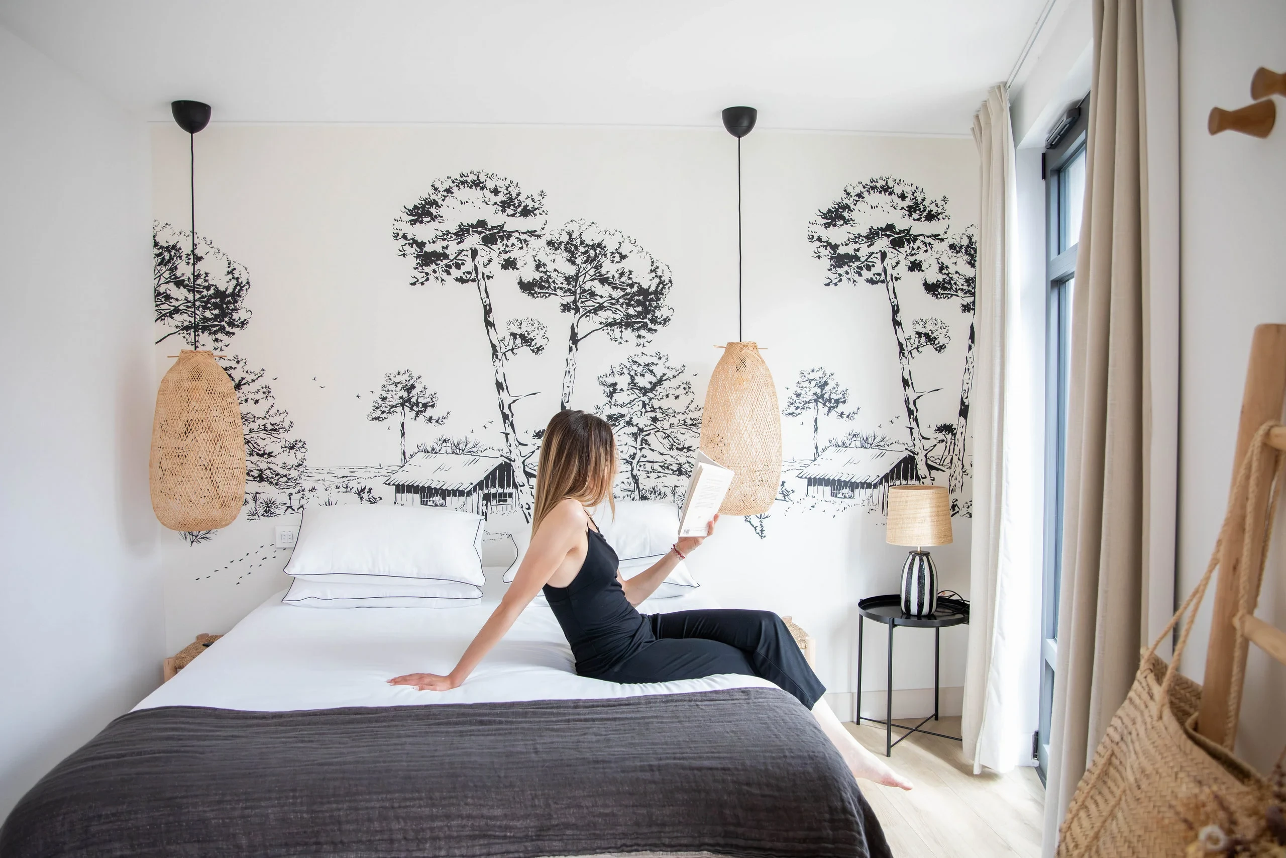 Femme assise sur le lit d'une chambre moderne avec fresque murale végétale - Appartement Vacances Perpignan - Les Suites Collection