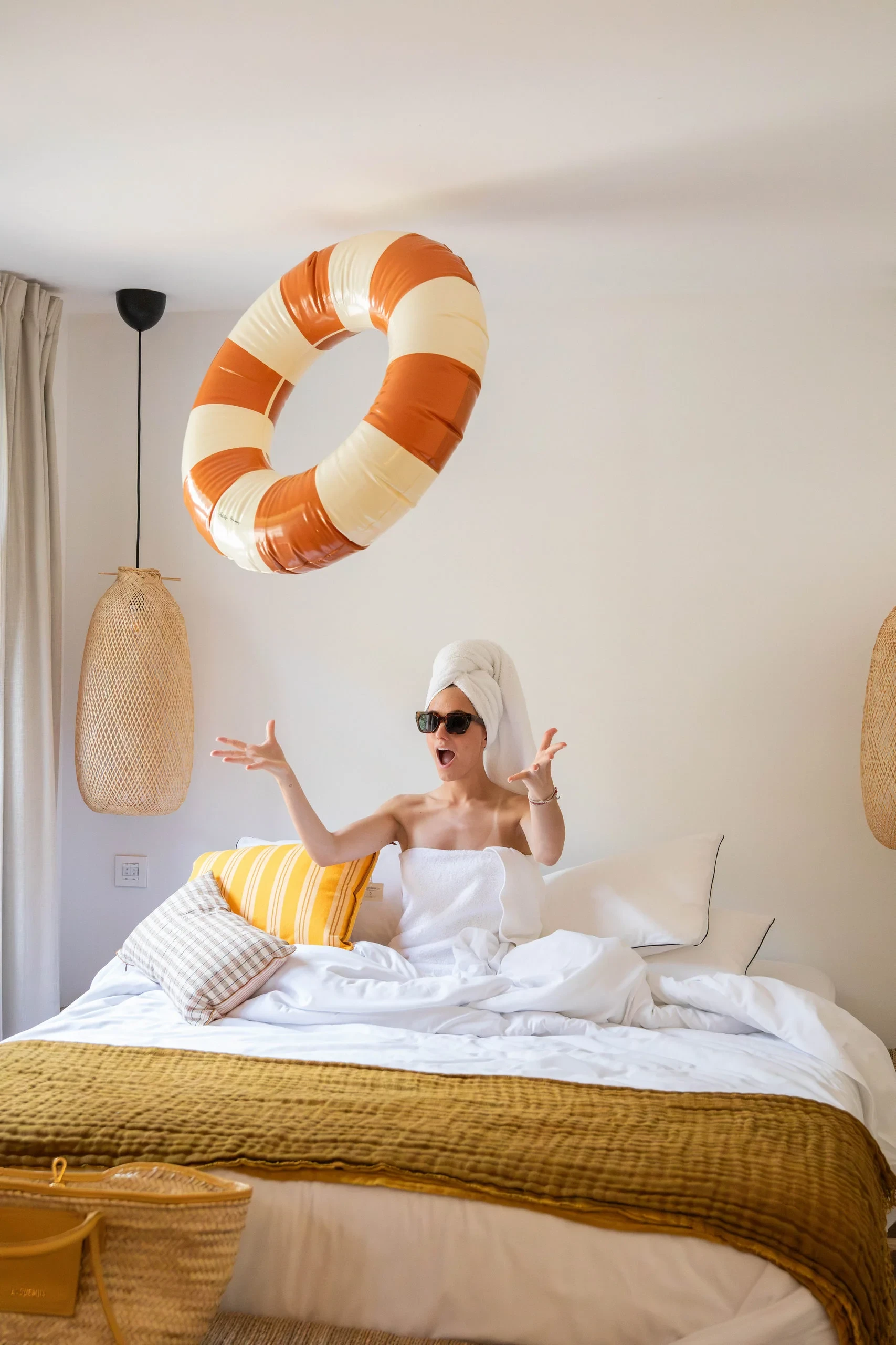 Femme assise sur le lit lançant une bouée rayée en l'air dans une chambre lumineuse - Vacances Collioure - Les Suites Collection